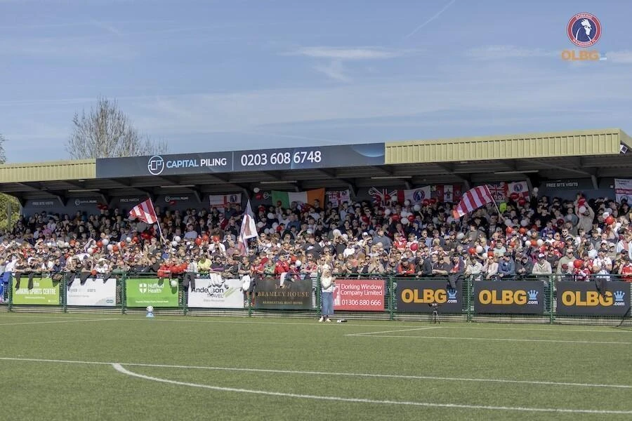 The home stand at meadowbank stadium, home of Dorking Wanderers FC, the stand is pcaked with fans in thier home red and white colours with flags flying